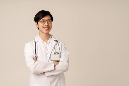 Image of asian young male doctor in medical white uniform smiling isolated over beige background