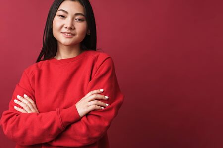 Close up of a pretty smiling young asian woman wearing casual clothes standing isolated over red background, arms foldedの写真素材