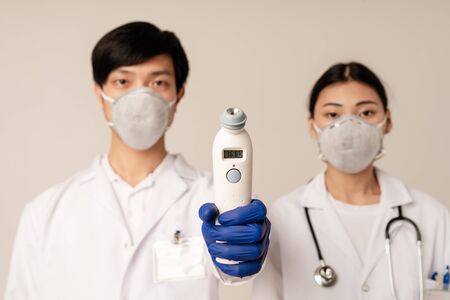 Image of asian young doctors wearing uniform and protective masks holding medical equipment isolated over beige backgroundの写真素材