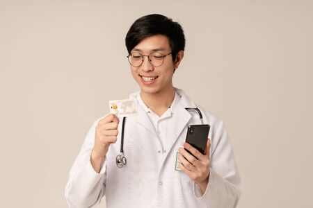 Image of asian young male doctor in medical white uniform holding credit card and cellphone isolated over beige backgroundの写真素材