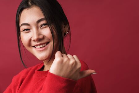 Close up of a pretty smiling young asian woman wearing casual clothes standing isolated over red background, pointing finger away at copy spaceの写真素材