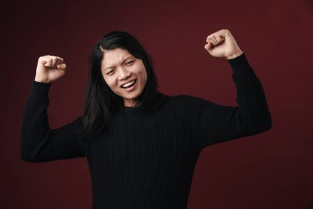 Portrait of a cheerful young asian woman standing isolated over dark background, wearing casual...