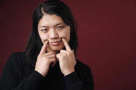 Image of sad asian man making fake smile with fingers isolated over burgundy background