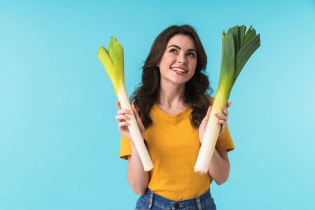 Photo of a dreaming positive young girl isolated over blue wall background holding leek.