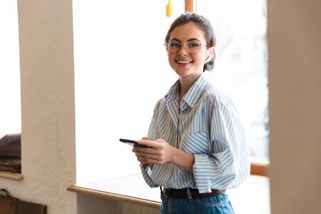 Attractive smiling young brunette businesswoman wearing wireless earphones on a call to a colleague shile standing at the window indoors, holding mobile phoneの写真素材