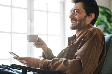 Image of handsome unshaven young man wearing eyeglasses drinking tea while resting in bright modern cafeの写真素材