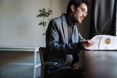 Image of handsome unshaven young businessman wearing suit examining documents in cafe