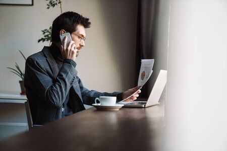 Image of handsome unshaven focused businessman examining documents and talking cellphone in cafe