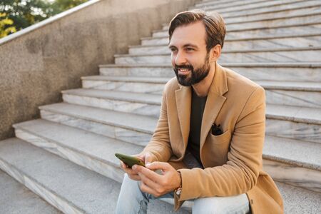 Image of handsome adult man in jacket holding smartphone while sitting on stairs outdoorsの写真素材