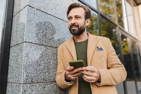 Image of handsome bearded adult man in sunglasses holding smartphone while walking on city street
