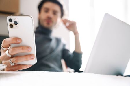 Image of handsome caucasian young man using laptop computer and cellphone while working in modern...