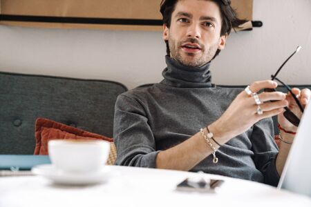 Image of handsome caucasian young man working on laptop computer and drinking coffee in modern cafe