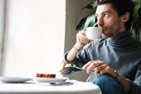 Image of handsome caucasian young man drinking coffee and eating dessert while having breakfast in bright cafeの写真素材