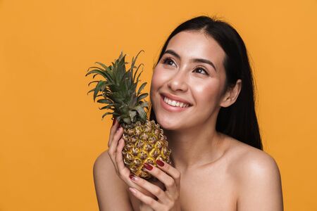 Beauty portrait of caucasian young brunette half-naked woman smiling and holding pineapple isolated over yellow backgroundの写真素材
