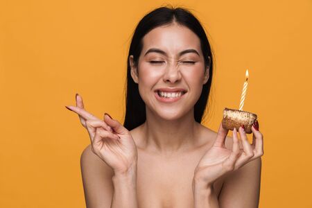 Beauty portrait of caucasian young brunette half-naked woman celebrating holiday and holding birthday cake isolated over yellow backgroundの写真素材