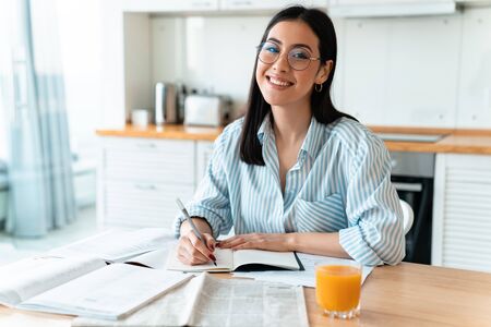 Image of a happy positive brunette young woman at the kitchen indoors at home writing notes studying.の写真素材