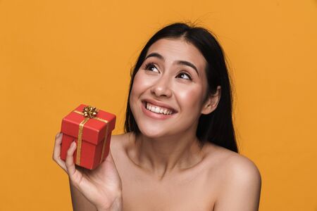 Beauty portrait of caucasian young brunette half-naked woman smiling while holding present box isolated over yellow backgroundの写真素材