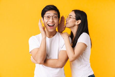 Image of smiling woman in eyeglasses whispering secret to surprised asian man isolated over yellow backgroundの写真素材