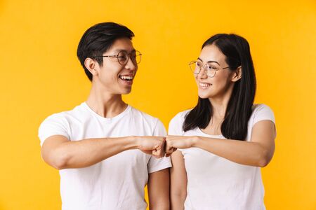 Portrait of a beautiful cheerful young asian couple wearing casual clothing standing isolated over yellow background, fist bumpingの写真素材