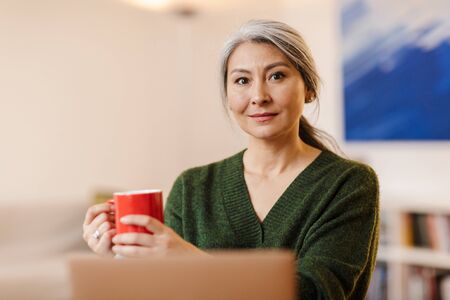 Image of positive pleased mature beautiful grey-haired woman using laptop computer indoors drinking...