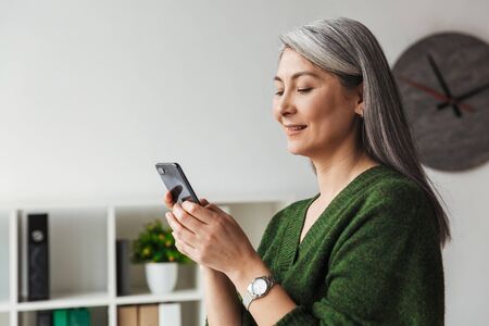 Photo of gray-haired smiling businesswoman typing on cellphone while working in officeの写真素材