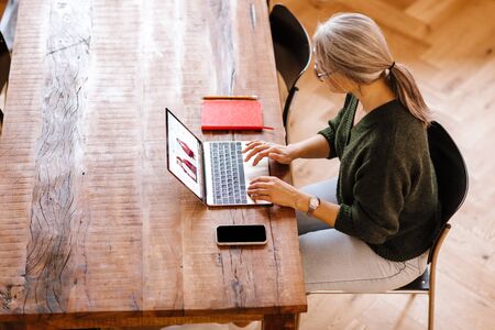 Photo of silver mature woman in eyeglasses using laptop while sitting at table in living roomの写真素材