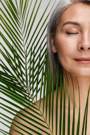 Beauty portrait of an attractive sensual mature topless woman with long gray hair standing isolated over gray background, eyes closed, posing with tropical leafの写真素材
