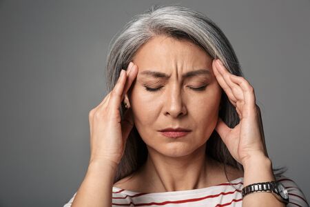 Image of adult mature tired woman wearing striped shirt touching her temples isolated over gray...