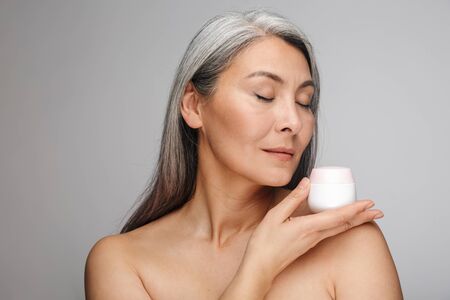 Beauty portrait of an attractive sensual mature topless woman with long gray hair standing isolated over gray background, eyes closed, showing cream containerの写真素材