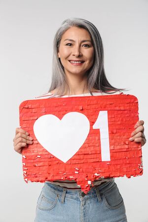 Image of adult mature happy woman with long white hair holding heart like symbol on placard isolated over gray backgroundの写真素材