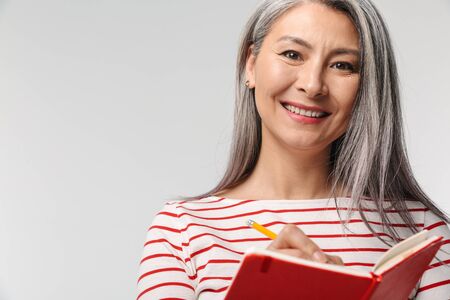Image of adult mature woman with long white hair holding diary book and pen isolated over gray backgroundの写真素材