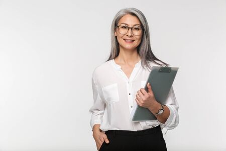 Portrait of an attractive smiling mature businesswoman in formal wear standing isolated over white background, holding a clipboardの写真素材