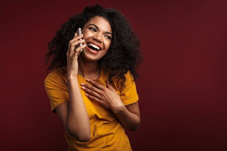 Image of beautiful brunette african american woman with curly hair smiling while talking on cellphone isolated over red backgroundの写真素材