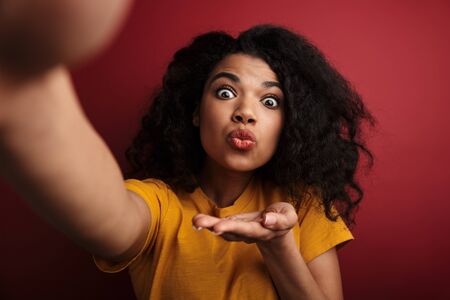 Image of beautiful brunette african american woman sending air kiss while taking selfie photo...
