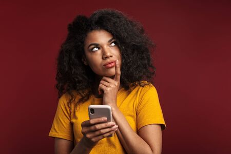 Image of beautiful brunette african american woman with curly hair looking upward and using cellphone isolated over red backgroundの写真素材