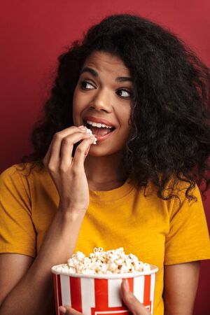 Image of beautiful brunette african american woman with curly hair smiling and holding popcorn bucket isolated over red backgroundの写真素材