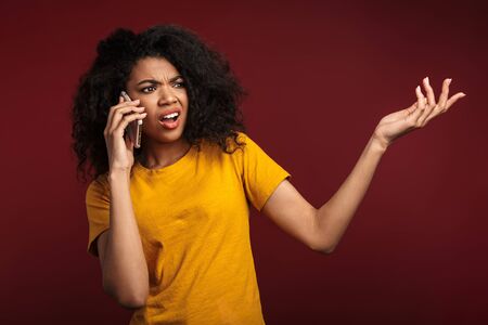 Image of displeased brunette african american woman with curly hair expressing outrage while talking on cellphone isolated over red backgroundの写真素材