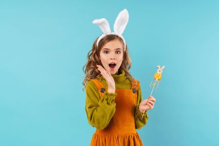Image of a cute shocked surprised young girl child in easter holiday bunny ears isolated over blue...