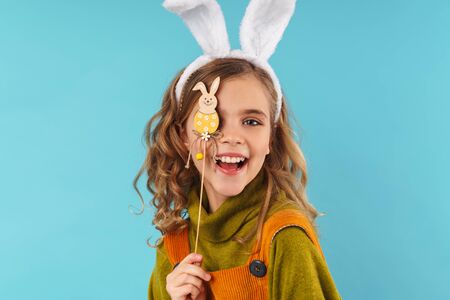 Photo of joyful curly girl in toy rabbit ears making with toy bunny and smiling isolated over blue backgroundの写真素材