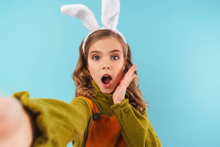 Photo of excited blonde girl in toy rabbit ears taking selfie photo and expressing surprise isolated...