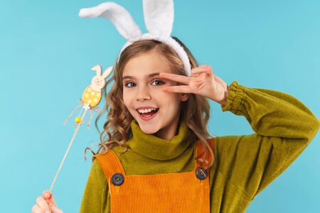 Photo of joyful curly girl in toy rabbit ears holding toy bunny and gesturing peace sign isolated...