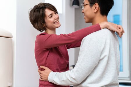 Photo of romantic multinational couple laughing and hugging while dancing in kitchen at home