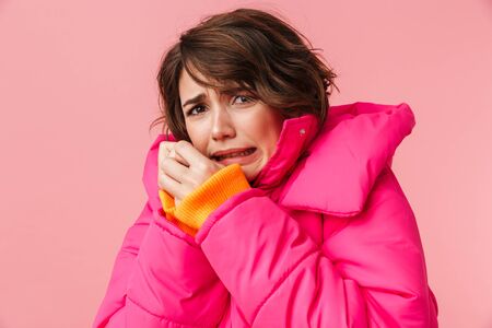 Portrait of young scared woman in warm coat looking at camera isolated over pink backgroundの写真素材