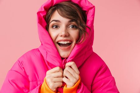 Portrait of young pleased woman in warm hood is freezing and smiling isolated over pink backgroundの写真素材