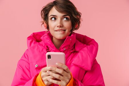 Portrait of young thinking woman in warm coat using headphones and cellphone isolated over pink backgroundの写真素材