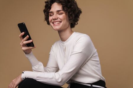 Image of laughing beautiful woman using cellphone while sitting isolated over beige background