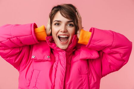 Portrait of young excited woman in warm coat grabbing her head and smiling isolated over pink...
