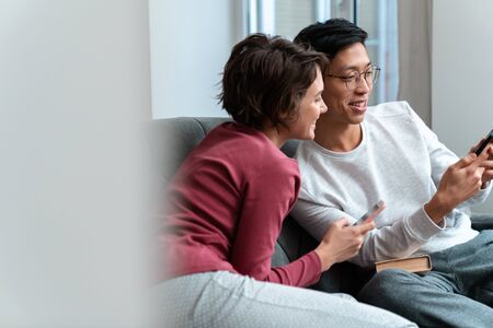 Photo of joyful multinational couple laughing and using smartphones while sitting on sofa at home