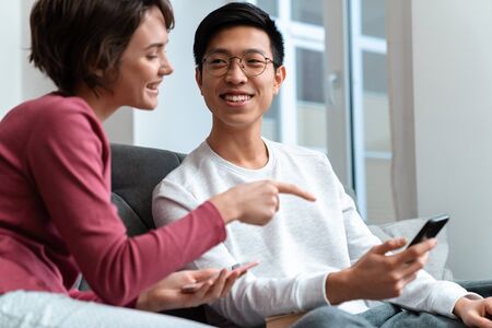 Photo of joyful multinational couple using and pointing finger at smartphone while sitting on sofa