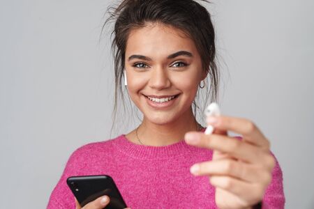 Image of cheerful beautiful woman using cellphone and wireless earphones isolated over gray...